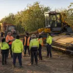 Workers attending a pre-start briefing beside parked almond harvesting machinery and a tractor-trailer